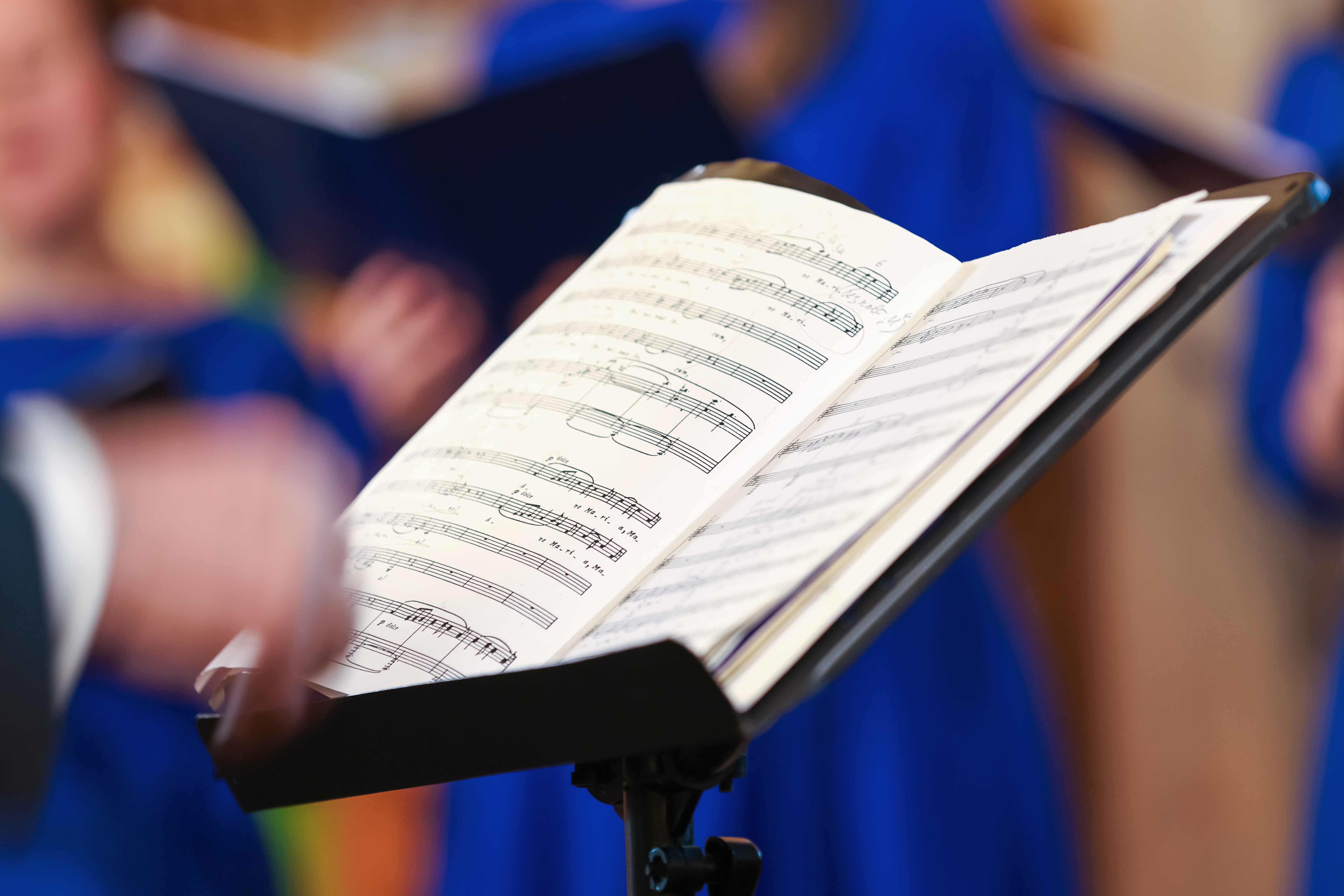 Open sheet music on a stand as a conductor leads a choir in blue robes, capturing focused singers, harmony and the intimate atmosphere of a choral rehearsal or live vocal performance